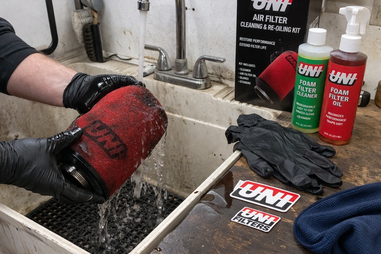 A person rinsing a red oiled foam performance air filter under a gentle stream of water over a utility sink. A filter cleaning and re-oiling kit is laid out on a workbench beside the dirty filter.