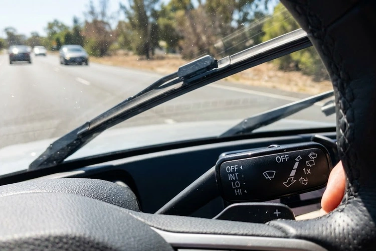 Close-up of car windshield wipers actively moving on a completely dry windshield in bright sunlight. The wiper stalk switch on the steering column is visible in the foreground.